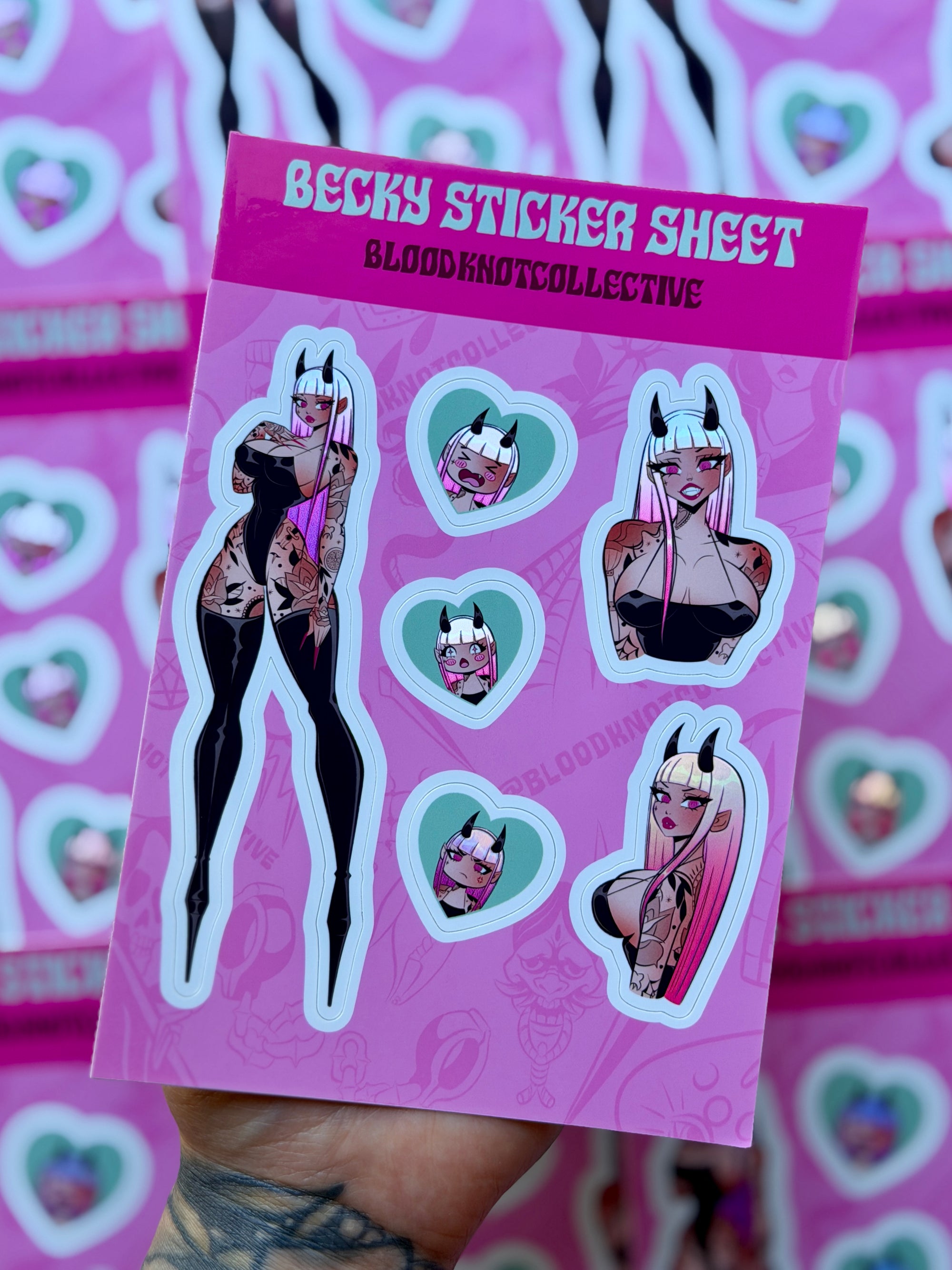 A hand holds a sticker sheet featuring an illustrated character with pink hair and horns, printed on a bright pink backing card. The card reads 'BECKY STICKER SHEET' and 'BLOODNOTCOLLECTIVE'.