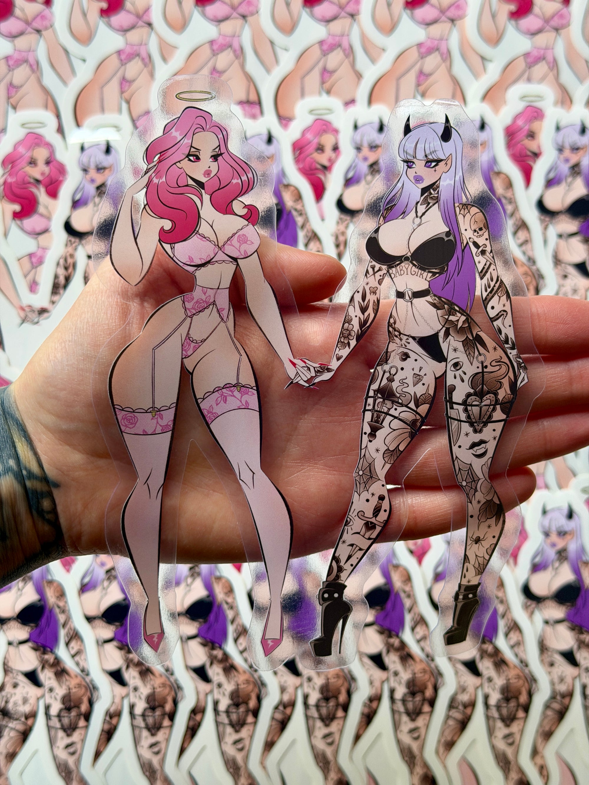 One die cut clear vinyl sticker of an angel with pink hair in lingerie holding hands with a tattooed devil with purple hair, with a heart between them. 