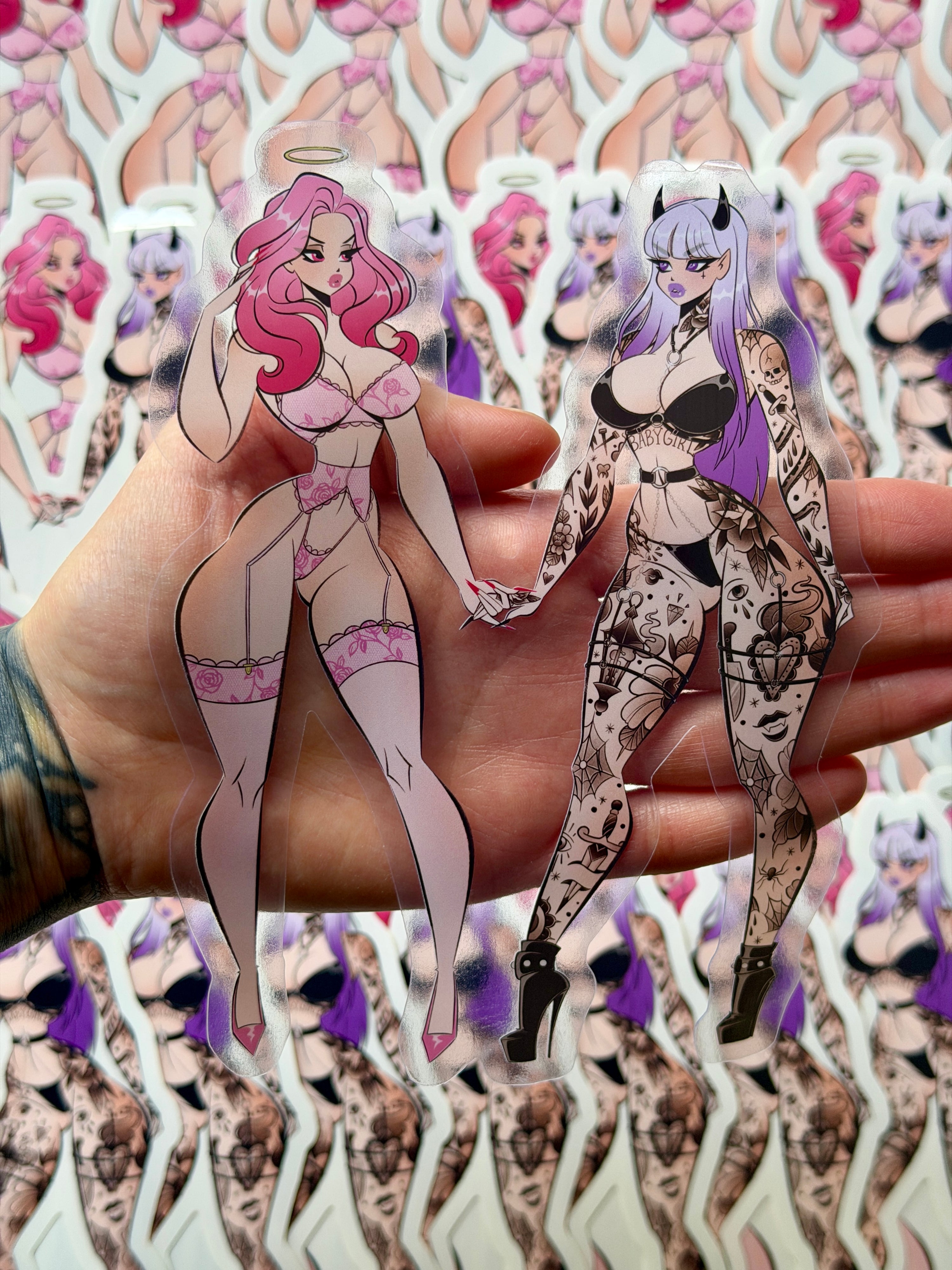 One die cut clear vinyl sticker of an angel with pink hair in lingerie holding hands with a tattooed devil with purple hair, with a heart between them. 